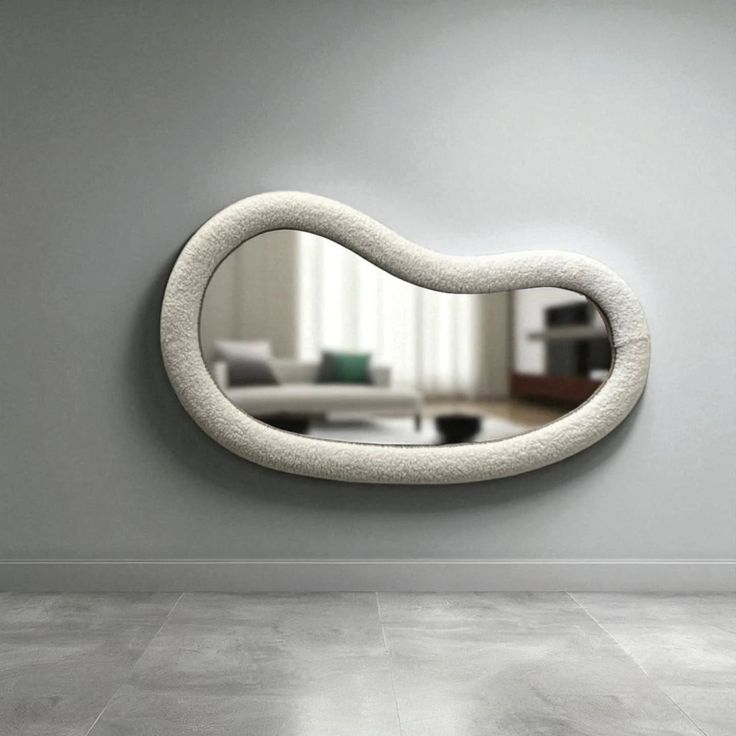 A large horizontal bean-shaped wall mirror with a textured cream bouclé frame mounted on a gray wall above concrete tile floors.