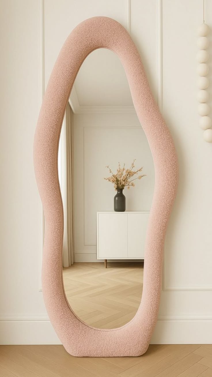 A tall, wavy full-length floor mirror with a textured blush pink bouclé fabric frame leaning against a paneled white wall on herringbone wood floors.