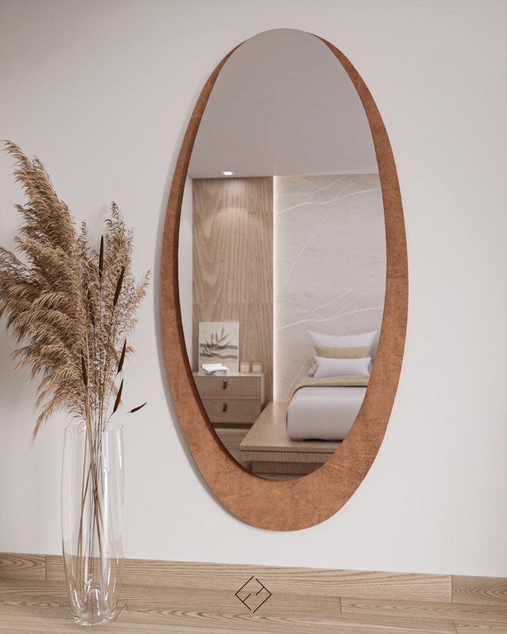 An elongated oval wall mirror with a wide-bordered terracotta rust fabric frame hung above a wooden floor next to pampas grass.