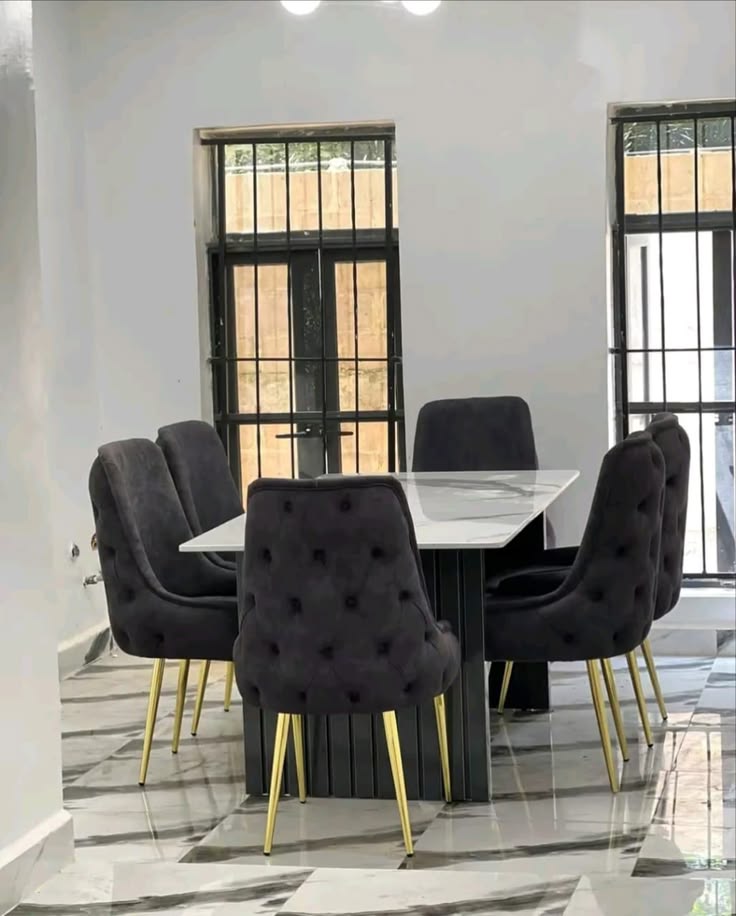 A contemporary dining area showcasing a rectangular white faux marble dining table with a fluted dark column base, paired with six deep charcoal gray button-tufted velvet chairs with polished gold legs on a gray marble-veined tile floor.