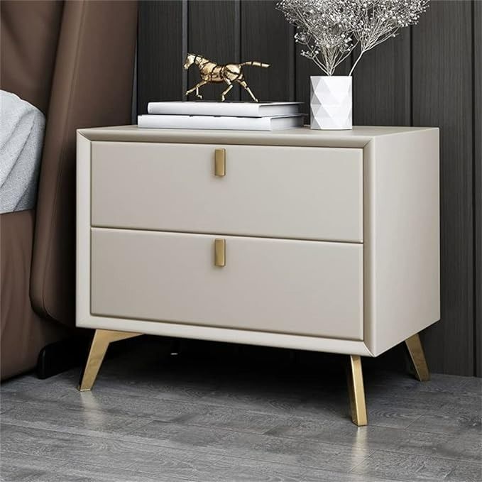 Modern off-white faux leather nightstand with two drawers and tapered gold legs standing next to a bed.