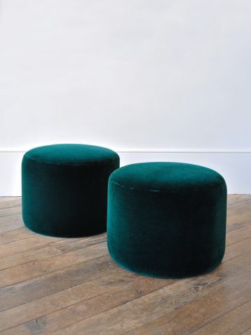 Two smooth, round emerald green velvet cylinder ottomans resting on a rustic wooden floor against a clean white wall, representing modern minimalist accent seating.