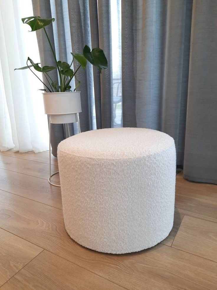 A solid cylindrical, cream-colored bouclé textured ottoman resting on a light wood floor, positioned next to a tall potted green plant with a white planter and stand, against a backdrop of grey and white curtains.