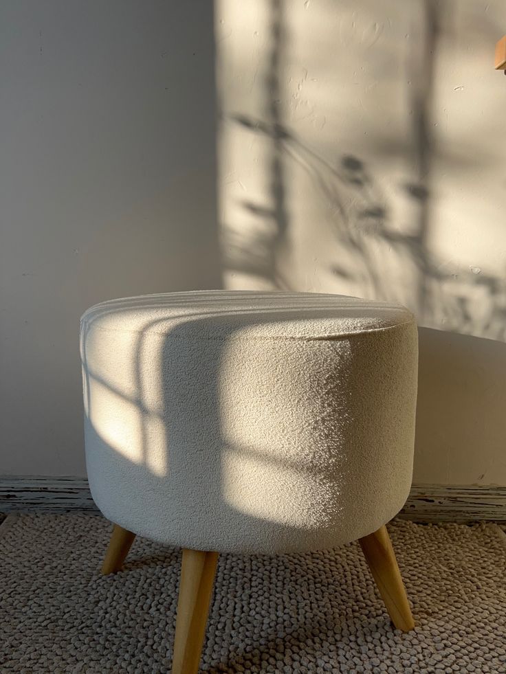 A small, round cream bouclé footstool ottoman with four light-wood splayed tapered legs, bathed in warm, geometric window sunlight on a textured rug against a plain white wall.