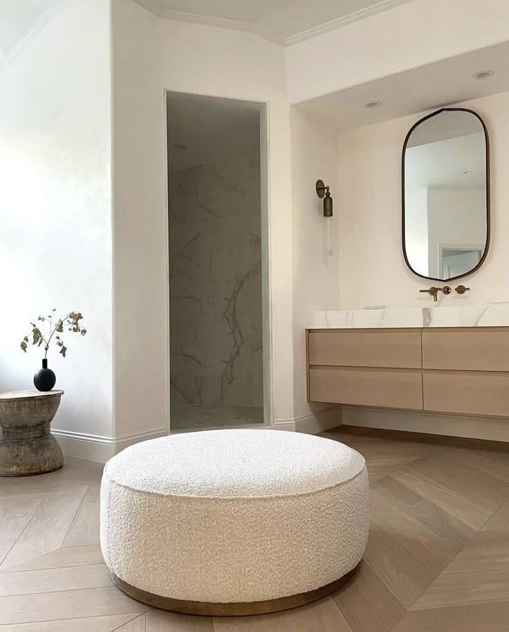 A large, round, cream-colored bouclé textured ottoman with a recessed brass metal base, positioned on light herringbone wood flooring in a serene, modern bathroom with a floating vanity.