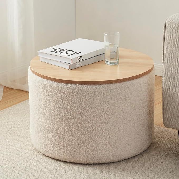 A round, cream-colored bouclé textured storage ottoman featuring a flat, light-wood tray top functioning as a coffee table, with a glass of water and a coffee table book placed on top.