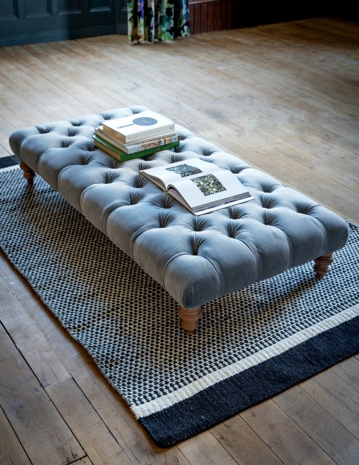A large rectangular dove gray velvet button-tufted coffee table ottoman styled with art books on a black and white patterned rug over rustic wood floors.