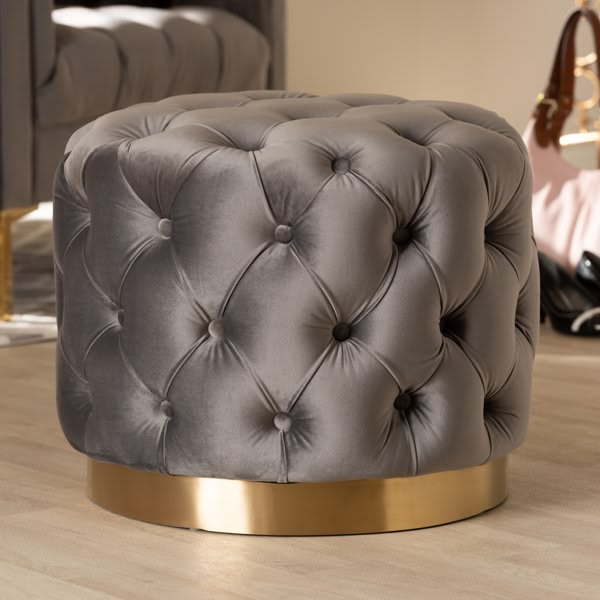A round, charcoal grey velvet ottoman with deep diamond button-tufting and a shiny gold metallic base, placed on a light wood floor with a handbag visible in the background.