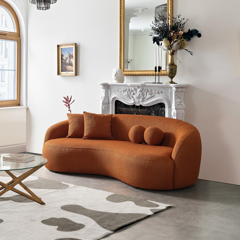 A contemporary curved bouclé sofa in terracotta burnt orange with two round ball pillows and two square pillows, styled in a living room next to a white fireplace mantel.