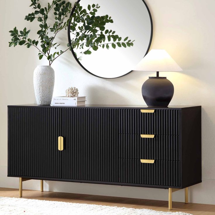 A contemporary matte black sideboard featuring vertically fluted panels, two cabinets, three drawers, and gold-toned metal legs styled against a wall with a round mirror and a lamp
