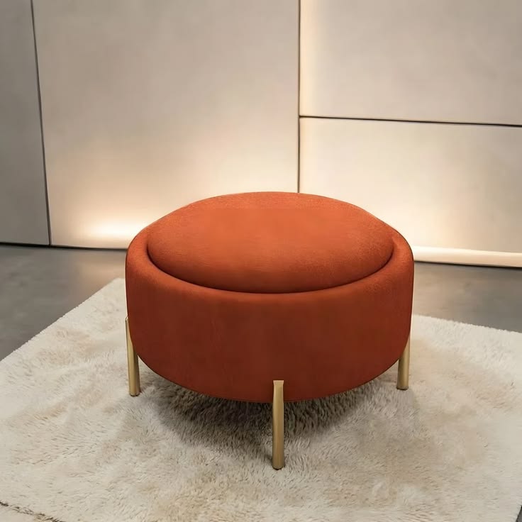 A round rust orange storage ottoman with a removable lid and cylindrical gold metal legs, sitting on a fluffy white shag rug in a room with modern white paneled walls.