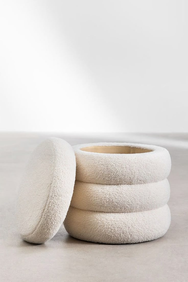 A cream-colored round boucle storage ottoman with a tiered ribbed exterior and a removable top lid, resting on a smooth grey concrete floor against a plain white wall.