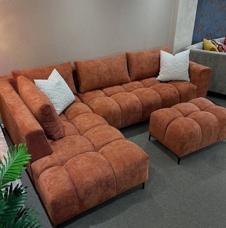 L-shaped terracotta chenille sectional sofa featuring deep biscuit tufting and a matching square ottoman styled with white textured throw pillows.